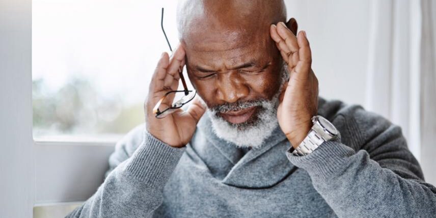 SeniorManWithMigraine-1135279293 (1) A man with a bald head holding his hands to his ears